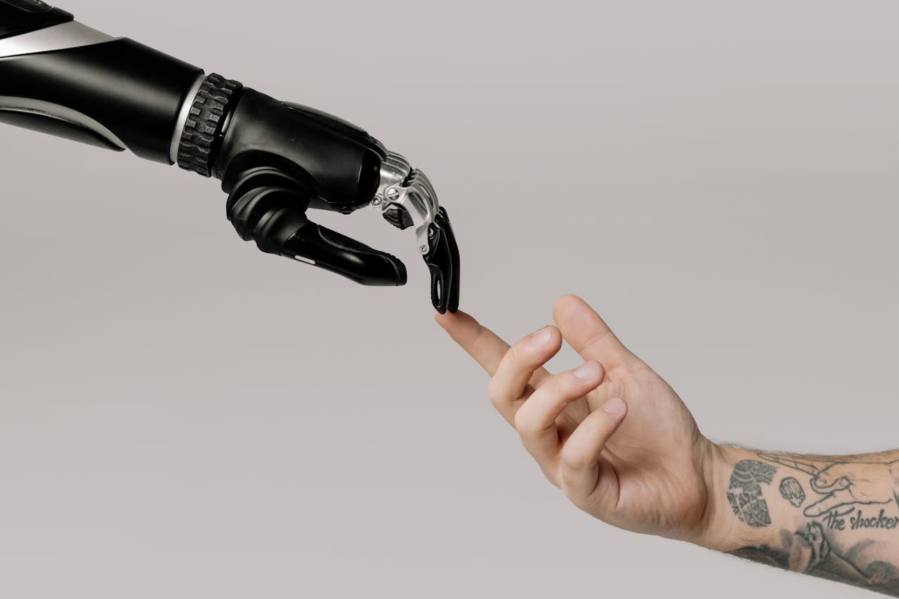 who-we-are A human hand with tattoos reaching out to a robotic hand on a white background.